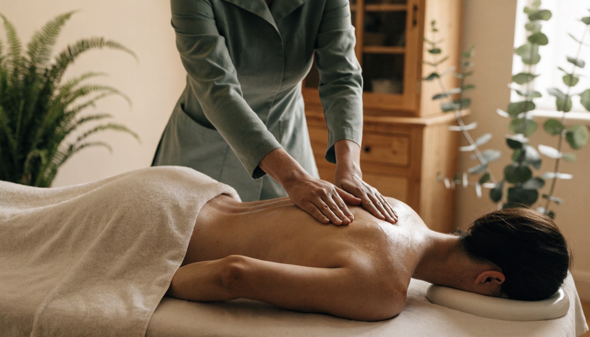Massage therapist in a sage-green uniform giving a back massage to a client lying face-down on a table, draped with a cream towel